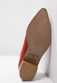 Suela de zapato de tacón alto de ante rojo con punta afilada, que presenta una suela de goma texturizada marrón y una base de tacón de madera. Nombre de la marca visible.