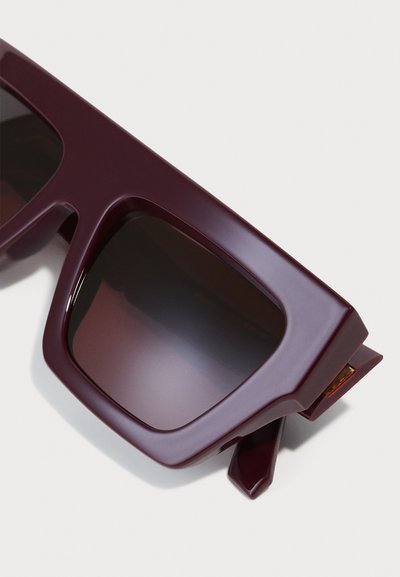 Lunettes de soleil maroon avec un design audacieux et angulaire. Comprend des verres teintés foncés et des accents pointus distinctifs sur les tempes. Matière en plastique.