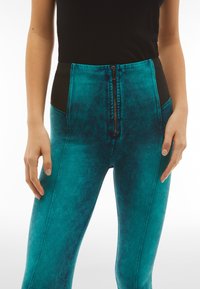 Dettaglio di leggings alta vita turchesi con pannelli laterali neri e una cerniera centrale frontale, indossati con un top senza maniche nero.