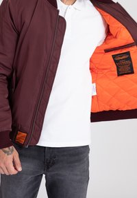 Vinsch bomberjacka med en vadderad orange foder. Har ribbade muddarna, dragkedjestängning och ett märke på fållen. Bärs över en vit polo-tröja.