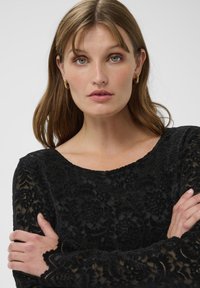 Top lung cu maneci lungi din dantelă neagră, având modele florale, margini scallopate la manșete și un decolteu larg. Tessut neted, texturat.