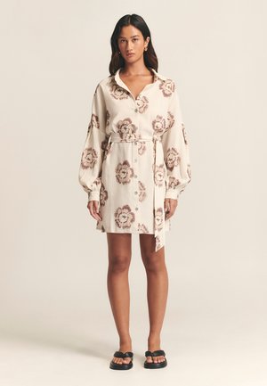 Langärmliges, knopfmloses Kleid in hellbeige mit floralem Muster in Brauntönen, mit einem taillierten Gürtel und entspannter Passform. Kombiniert mit schwarzen Rutschsandalen.