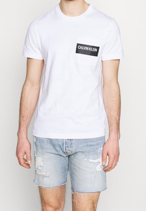 Biała bawełniana koszulka z czarną prostokątną kieszenią z logo "CALVIN KLEIN". Połączona z postrzępionymi, jasnoniebieskimi szortami dżinsowymi.