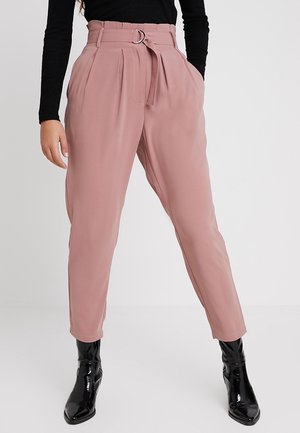 Mujer con pantalones ajustados de talle alto en color rosa polvo, con cinturón a juego y botines negros, posando frente a un fondo claro y liso.