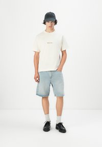 Tricou bej deschis cu detalii brodate, pantaloni șort din denim și șapcă neagră. Pantofi negri cu tălpi texturate, șosete albe vizibile deasupra pantofilor.
