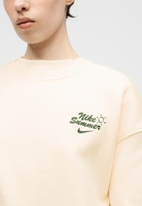 Μπεζ φούτερ με πράσινη επιγραφή "Nike Summer", ένα μικρό εικονίδιο ήλιου και το λογότυπο Nike στην αριστερή πλευρά του στήθους, φορεμένο από άτομο με κοντά μαλλιά.