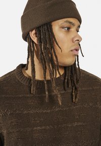 Maglione marrone a maglia con motivi testurizzati e colletto a coste, abbinato a un berretto a coste marrone coordinato. Dreadlocks scendono sotto il cappello.