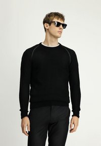 Czarny sweter dzianinowy w wzór rybiej ości, z prążkowanymi mankietami i dołem. Noszony na białej koszulce z okrągłym dekoltem, zestawiony z ciemnymi spodniami.