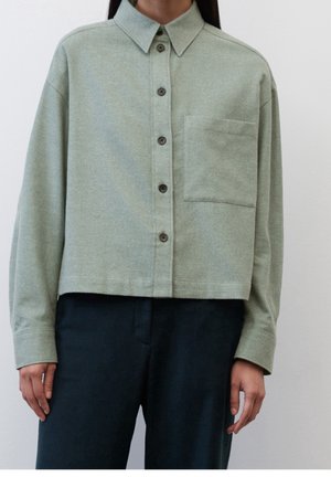 Femme portant une chemise courte boutonnée vert clair avec une poche poitrine et des manches longues, associée à un pantalon foncé.