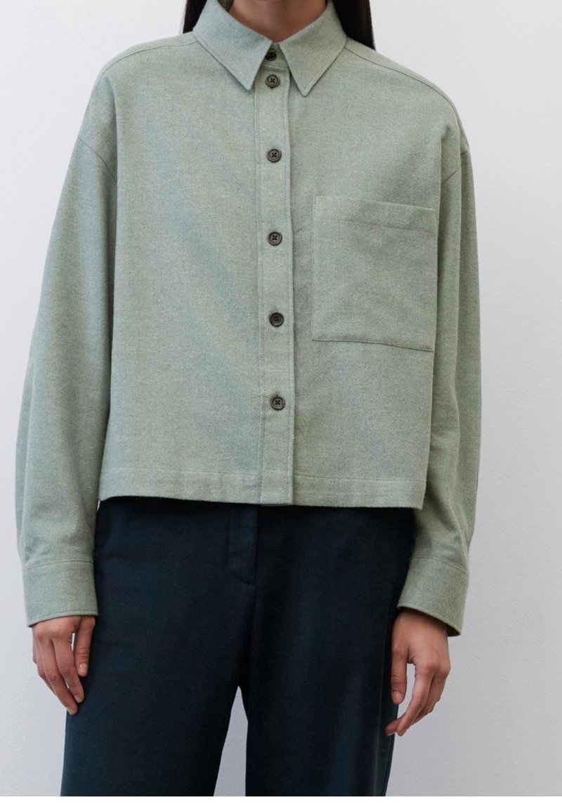 Femme portant une chemise courte boutonnée vert clair avec une poche poitrine et des manches longues, associée à un pantalon foncé.