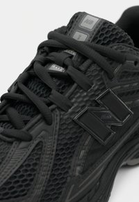 Chaussure de sport noire avec un dessus en maille texturée, des lacets plats noirs et une semelle en caoutchouc. Présente un logo noir bien visible et des accents réfléchissants.