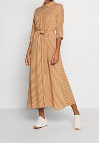 Vestito camicia a righe marroni e bianche, lunghezza midi, davanti con bottoni, maniche a tre quarti, vita stretta con cintura annodata, abbinato a scarpe da ginnastica bianche.