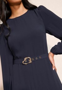 Jūras tērps ar garām piedurknēm, apaļu izgriezumu un pieguļošu jostasvietu, kurai ir teksturēts zelta sprādze un atbilstoši grometi.