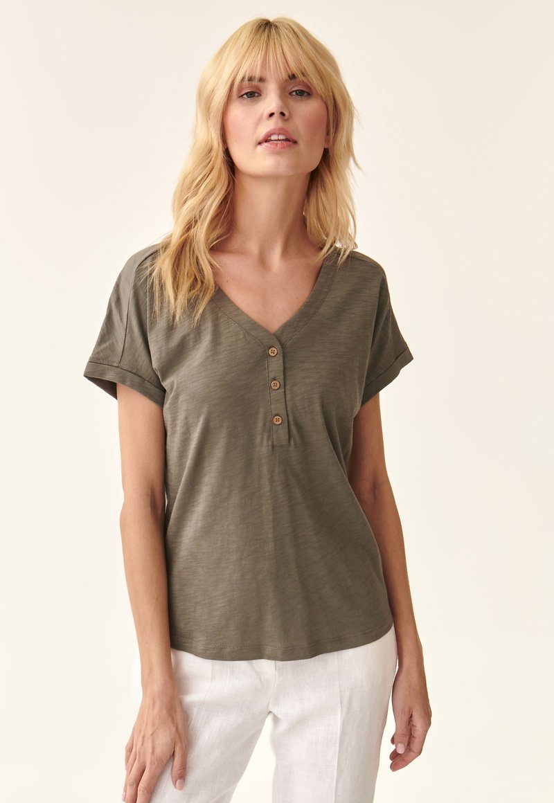 Olivgrünes V-Ausschnitt-T-Shirt aus weichem Stoff, mit umgeschlagenen Ärmeln und drei Holzknöpfen am Kragen. Kombiniert mit weißen Leinenhosen.