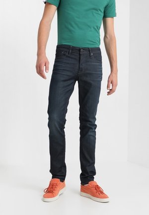 Mężczyzna ubrany w ciemnoniebieskie obcisłe dżinsy, zieloną koszulkę z krótkim rękawem oraz pomarańczowe tenisówki, stojący na białym tle.