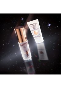 Spray fissante Airbrush Flawless con cappuccio oro rosa; e primer invisibile UV Poreless con SPF 50 in un tubo bianco. Entrambi esposti su uno sfondo scuro.