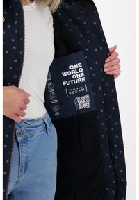 Giacca navy con piccoli punti blu, chiusura con zip e un morbido rivestimento interno; l'etichetta indica materiali vegani approvati da PETA.