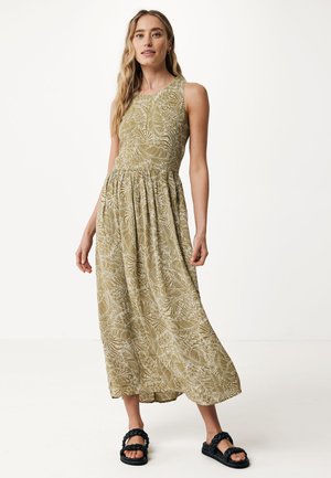 Groene, gemusterde maxi-jurk met mouwloos ontwerp en een gerimpelde taille. Fijn gedetailleerd met bladvormen in een lichte kleur. Draagt zwarte sandalen.