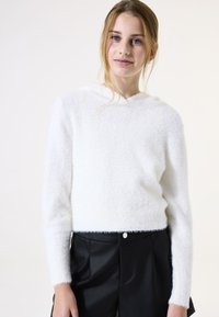 Weißer, flauschiger, kurz geschnittener Pullover mit Kapuze, langen Ärmeln und strukturiertem Stoff. Kombiniert mit schwarzen, hochgeschnittenen Hosen aus Kunstleder.