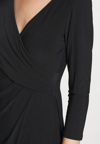 Svart wrap-klänning med trekvartsärmar, mjukt tyg, v-ringning och plisserade detaljer, med en figursydd midja och flödande kjol.