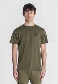 Camiseta de algodón verde oliva con cuello redondo, manga corta y un pequeño parche de logo negro en el lado izquierdo del pecho. Textura suave.