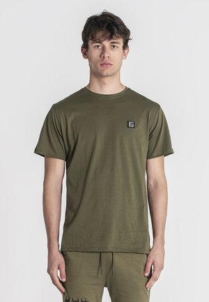 Olijfgroen katoenen T-shirt met een ronde hals, korte mouwen en een klein zwart logopatched op de linkerborst. Soepele textuur.