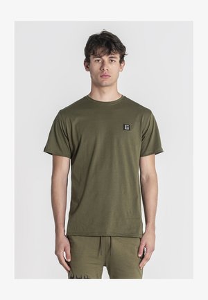 Olijfgroen katoenen T-shirt met een ronde hals, korte mouwen en een klein zwart logopatched op de linkerborst. Soepele textuur.