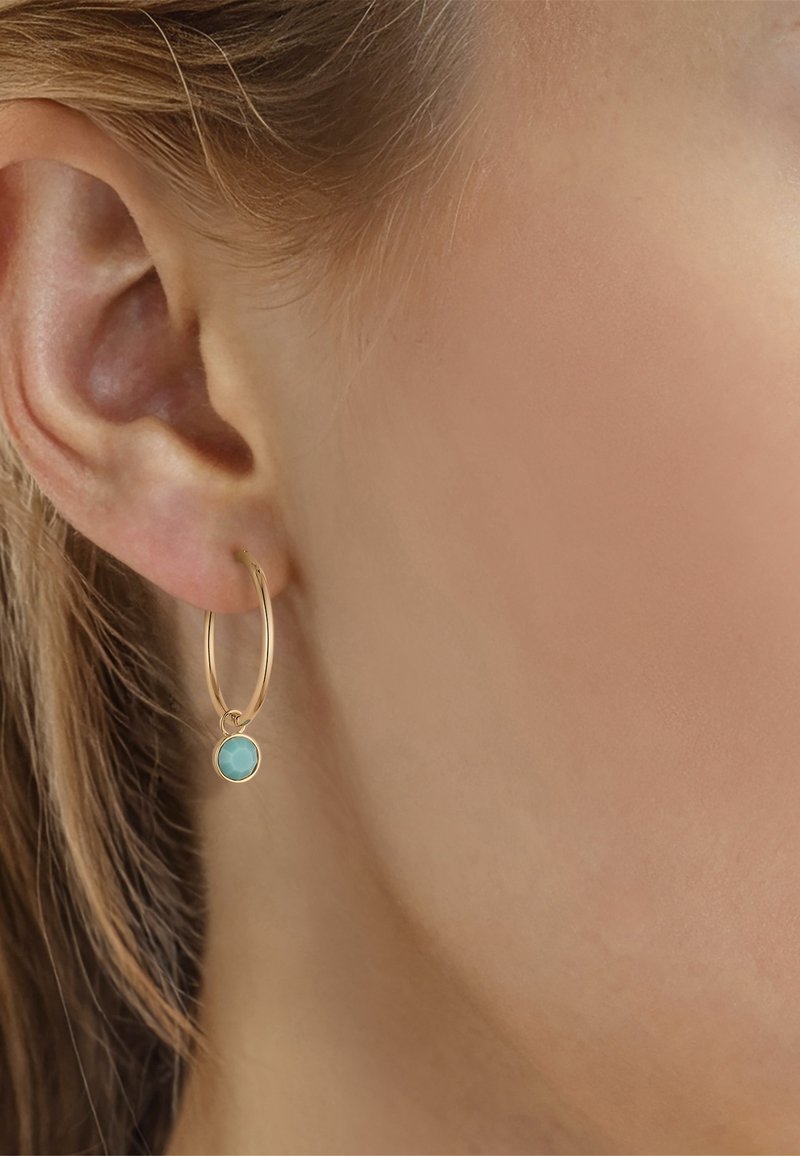 Primer plano de la oreja de una mujer con un pendiente de aro dorado con un pequeño colgante redondo de turquesa.