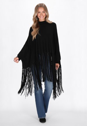 Poncho nero realizzato in tessuto morbido, con frange lunghe all'orlo, un collo alto e maniche ampie. Indossato sopra jeans blu e stivali neri.