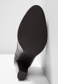 Zapato de tacón alto de cuero negro con suela de goma texturizada. El tacón es delgado y angular, mientras que la parte superior presenta un acabado suave.