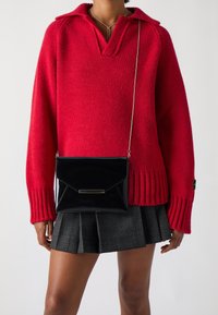 Červený pletený svetr s výstřihem do V a žebrovanými manžetami, doplněný černou patentní křížovou kabelkou s klopou a kovovým akcentem.