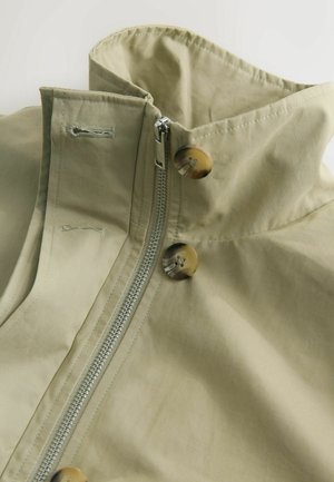 Helle Khaki-Jacke mit hohem Kragen, ausgestattet mit einem Reißverschluss und zwei großen braunen Knöpfen. Hergestellt aus glattem Stoff mit weicher Textur.