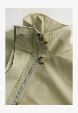 Helepruun khaki jakk kõrge kraega, millel on lukk ja kaks suurt pruuni nuppu. Valmistatud siledast kangast pehme tekstuuriga.