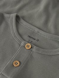 Primer plano del escote de una camisa de tela gris acanalada con dos botones marrones claros y etiqueta de ropa visible dentro del cuello.