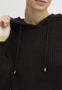 Svart ribbad huvtröja med dragsko i huvan, med metalliska aglets. Tyget har en strukturerad yta och en avslappnad passform.