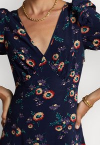 Vestido navy con estampado floral en rojo, crema y púrpura. Cuenta con un escote en V profundo, mangas abullonadas y detalles fruncidos en la cintura. Collar de cadena dorada.