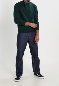 Mörkgrön sweatshirt med ribbstickade muddar, parat med blå denimjeans med kontrasterande sömmar. Svarta sneakers med vita sulor full ändar looken.