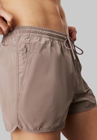 Pantaloni da bagno leggeri e marroni con cintura elastica, cordoncino regolabile e tasche laterali. Texture liscia con una finitura leggermente lucida.