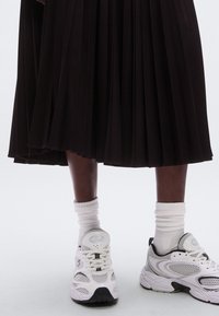 Jupe plissée noire tombant au-dessus de la cheville, associée à des chaussures de sport blanches et des chaussettes blanches côtelées. Les chaussures présentent des détails en mesh et des accents noirs.