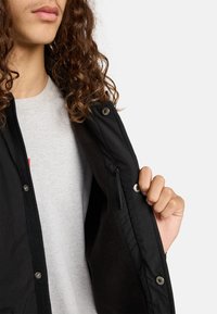 Zwarte jas met een ritsvak en drukknoopsluitingen. Zachte, zwarte fleece voering zichtbaar. Model draagt eronder een grijs T-shirt.