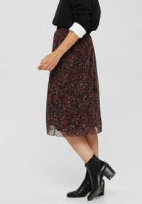 Personne portant un pull noir, une jupe fleurie longueur genou avec des fleurs rouges et violettes, et des bottines noires à talons, debout de côté.