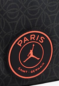 Czarna tkanina z tonalnym wzorem geometrycznym; posiada okrągłe logo z gumy w kolorze pomarańczowym i czarnym, z napisem "PARIS" i "SAINT-GERMAIN".