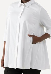 Camicia bianca con bottoni, colletto, maniche a 3/4 e tasche laterali. Caratterizzata da una vestibilità ampia e un tessuto morbido e testurizzato.