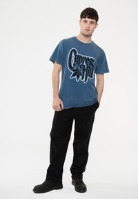Blue cotton t-shirt featuring a black graphic design with "Cypress Hill" text. Paired with black loose-fitting pants and black shoes.