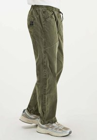 Persoon in olijfgroene cargobroek met elastische tailleband en beige-grijze sneakers, zijwaarts staand op een effen witte achtergrond.