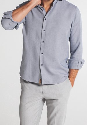 Hombre que lleva una camisa de botones texturizada de color azul claro con las mangas remangadas y pantalones gris claro, con la mano en el bolsillo y tocándose el cuello.
