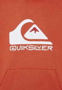 Logo Quiksilver bianco con un grafico di onda e montagna sopra il nome del marchio su uno sfondo di tessuto arancione con una tasca frontale.