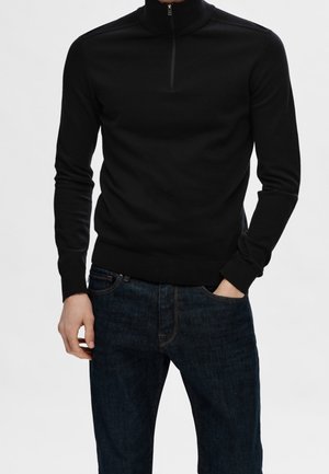 Jersey de punto negro de cuello alto con media cremallera, combinado con unos vaqueros oscuros que tienen bolsillos estándar y un sutil pespunte azul.