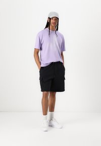 T-shirt de manga curta lilás clara, calções cargo pretos, boné cinzento, ténis brancos, em pé contra um fundo branco, com as mãos nos bolsos.