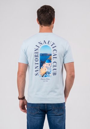 Bărbat purtând un tricou bleu deschis cu grafică a unui sat de coastă din Santorini și o cupolă albastră, asortat cu blugi albaștri, stând cu fața spre spate pe fundal alb.
